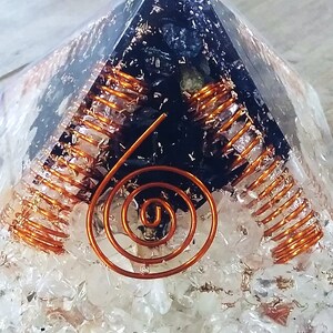 May include: A clear quartz pyramid with a black crystal inside. The crystal is surrounded by copper wire coils. The pyramid is sitting on a wooden surface.
