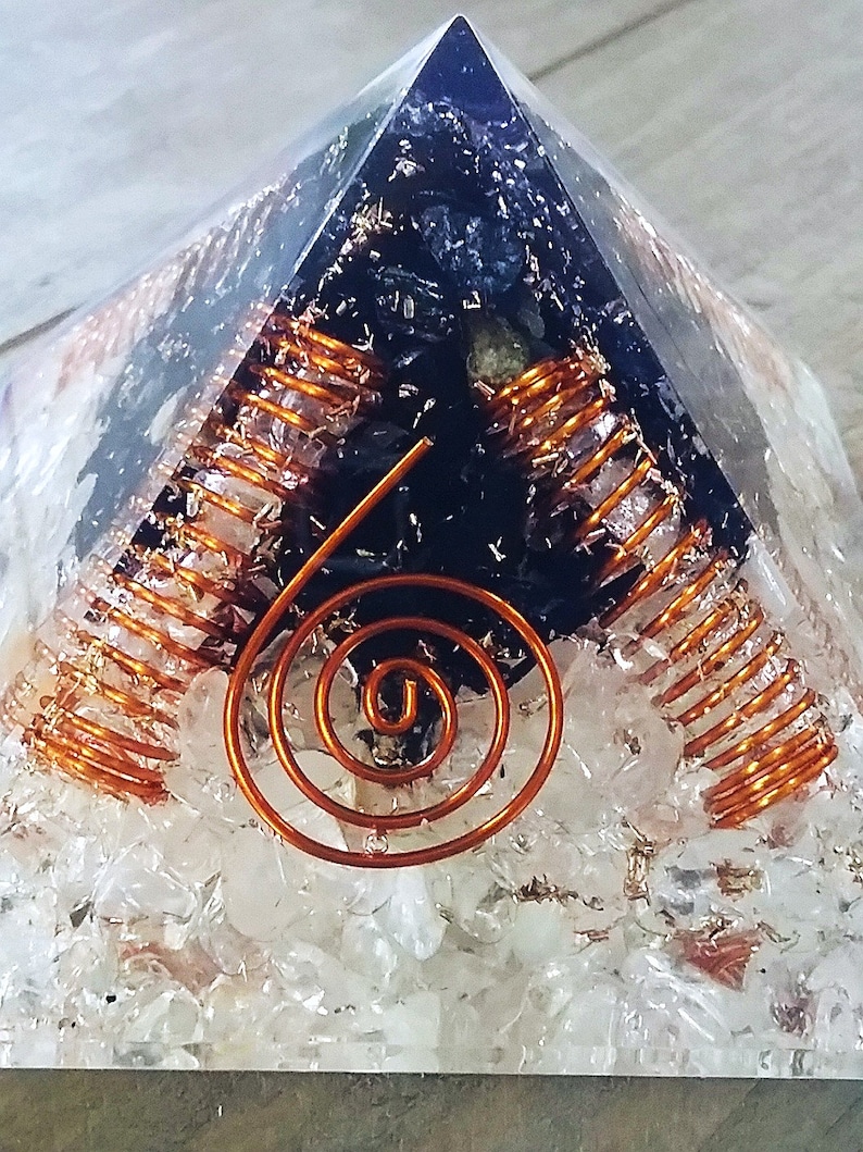 May include: A clear quartz pyramid with a black crystal inside. The crystal is surrounded by copper wire coils. The pyramid is sitting on a wooden surface.