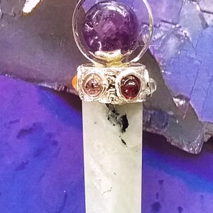 May include: A silver pendant with a clear crystal point and a round amethyst stone in a silver cage. The pendant is on a black cord and is surrounded by a purple background.
