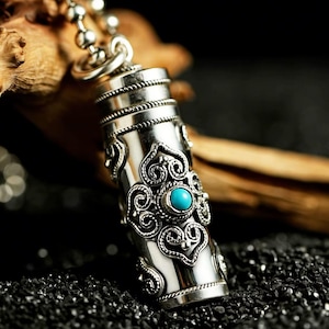 May include: A cylindrical silver pendant on a ball chain featuring intricate metal scrollwork and a single round turquoise stone set in the center. The metal surface is polished and reflective, showcasing detailed filigree patterns.