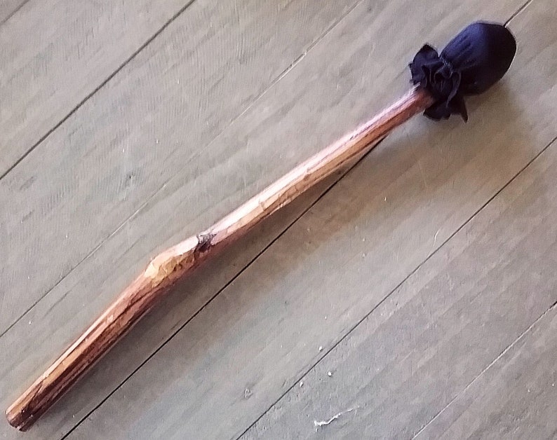 May include: A long, slender wooden wand featuring a natural, light brown bark texture and a smooth, polished finish. At one end, a dark navy blue fabric pouch is gathered and secured with a matching fabric tie, creating a rounded, bulbous tip.