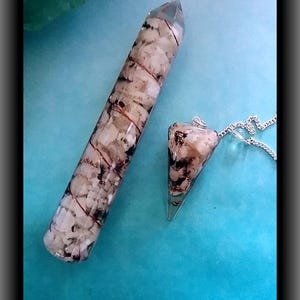 Op de afbeelding: Een heldere kristallen punt van hars en een bijpassende pendel. Beide zijn gevuld met witte en bruine stenen en omwikkeld met koperdraad. De pendel hangt aan een zilveren ketting. De items worden gepresenteerd op een turquoise achtergrond.