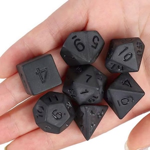 May include: A set of seven black polyhedral dice, each with white numbering, held in a hand. The dice include a four-sided die, a six-sided die, an eight-sided die, a ten-sided die, a twelve-sided die, a twenty-sided die, and a percentile die.