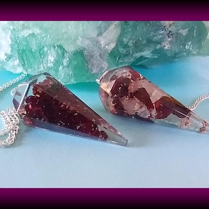 May include: Two red and clear crystal dowsing pendulums with silver chains. The pendulums are on a light blue surface.