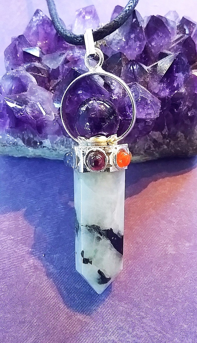 May include: A silver pendant with a clear crystal point, surrounded by a circle of amethyst and four smaller stones in red, orange, blue, and purple. The pendant is hanging from a black cord.
