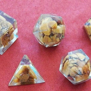 May include: Five clear resin dice with brown and gold flecks inside. The dice are a variety of shapes, including a cube, a pyramid, and a dodecahedron.
