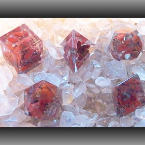 May include: Five red gemstone dice, each with a different shape, are displayed on a bed of clear quartz crystals. The dice are made of clear resin with red gemstone inclusions.