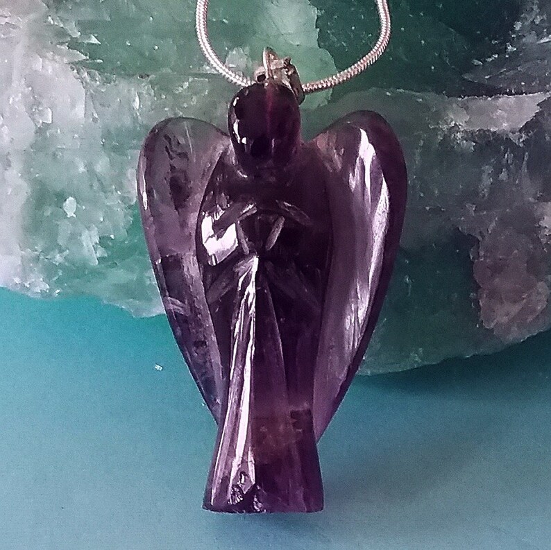 May include: A purple amethyst angel pendant carved from a single piece of crystal. The angel is wearing a silver chain.