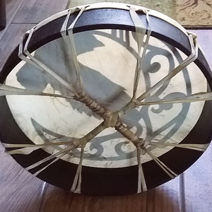 May include: A circular frame drum with a dark brown wooden rim and a light-colored drumhead. The drumhead features a dark gray graphic design and is secured to the frame with light-colored lacing that crisscrosses in the center of the back.
