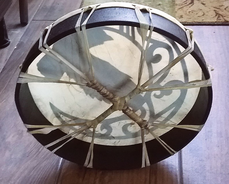 May include: A circular frame drum with a dark brown wooden rim and a light-colored drumhead. The drumhead features a dark gray graphic design and is secured to the frame with light-colored lacing that crisscrosses in the center of the back.