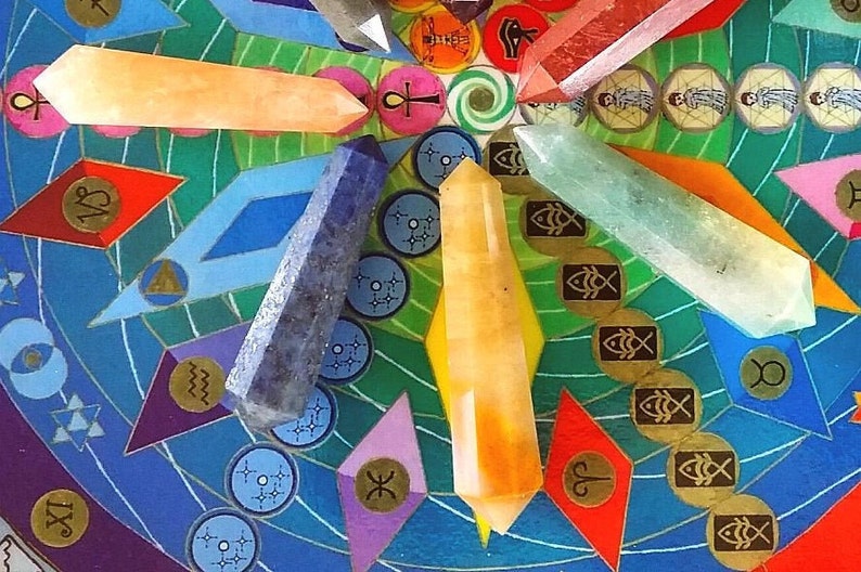 May include: Five polished crystal points in orange, dark blue, yellow, clear, and reddish-pink arranged on a circular board featuring colorful geometric patterns, zodiac symbols, and various icons including an ankh and fish.