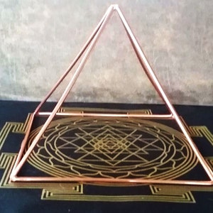 K&ouml;nnte beinhalten: Ein Pyramidenrahmen aus Kupferdraht auf einer schwarzen Oberfl&auml;che mit einem goldenen geometrischen Muster. Die Pyramide besteht aus d&uuml;nnem Kupferdraht und hat eine quadratische Basis.