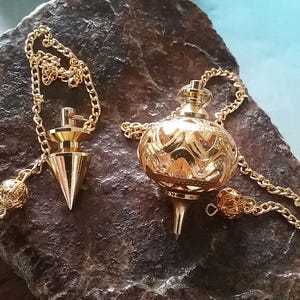 Pode incluir: Dois pêndulos dourados com correntes repousam sobre uma superfície escura e texturizada. Um é cónico, o outro redondo e com padrão. Ambos têm correntes douradas e detalhes decorativos.