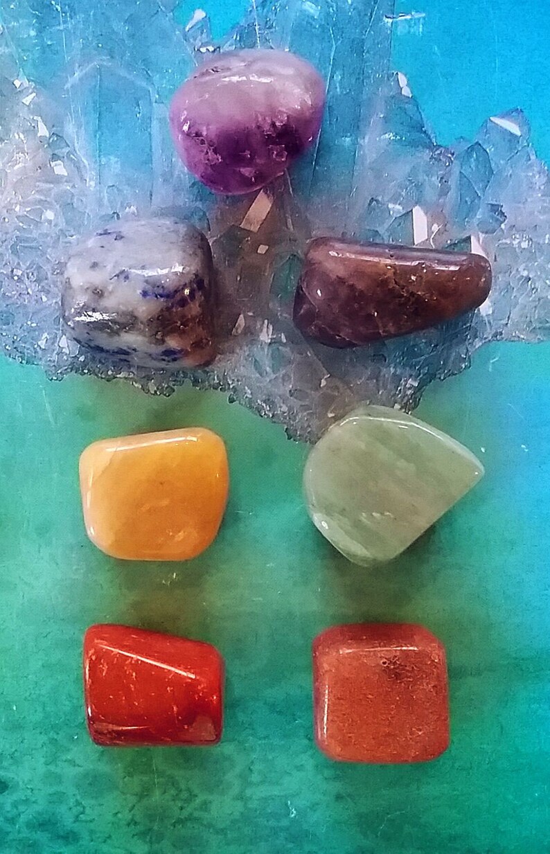 May include: Seven tumbled stones of various colours and shapes, including purple, blue, brown, yellow, green, and orange. The stones are arranged on a blue and green background.