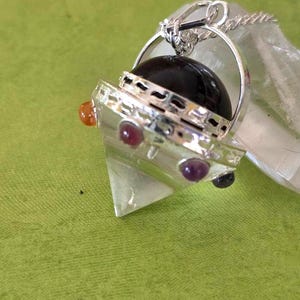 May include: A silver-toned pendant featuring a clear, triangular-shaped crystal base topped with a smooth, dark, rounded stone. The metal setting is adorned with small, colorful, circular cabochon gemstones in orange, red, and purple hues.