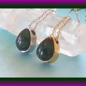 May include: Two teardrop-shaped pendants with green gemstone inlays. One pendant is on a silver chain and the other is on a gold chain. Both pendants are resting on a white crystal.