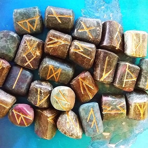 May include: A collection of 18 small, irregularly shaped stones with gold runes carved into their surfaces. The stones are a mix of brown, green, and red.