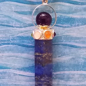 Puede incluir: Un colgante de plata con una piedra preciosa de color p&uacute;rpura oscuro en la parte superior, rodeado por un anillo de plata. El colgante tiene una piedra preciosa larga, puntiaguda, azul y dorada unida a la parte inferior.