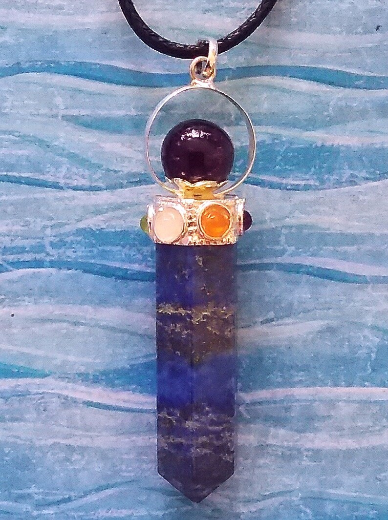 Puede incluir: Un colgante de plata con una piedra preciosa de color p&uacute;rpura oscuro en la parte superior, rodeado por un anillo de plata. El colgante tiene una piedra preciosa larga, puntiaguda, azul y dorada unida a la parte inferior.