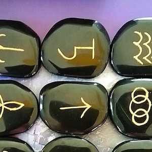 May include: Set of ten black obsidian stones with gold symbols. The symbols include an arrow, a circle with a dot in the center, a circle with three dots, a circle with two dots, a circle with a line through it, a circle with a cross in it, a circle with a triangle in it, a circle with a square in it, a circle with a star in it, and a circle with a crescent moon in it.