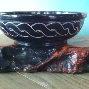 May include: A black slate bowl with a carved white Celtic knot design. The bowl is sitting on a wooden stand.