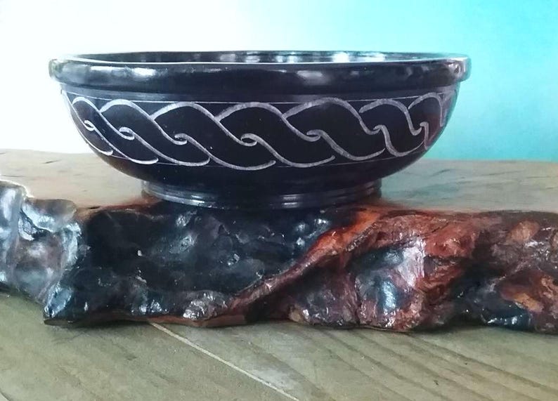 May include: A black slate bowl with a carved white Celtic knot design. The bowl is sitting on a wooden stand.