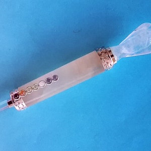May include: A crystal wand featuring a carved angel at one end and a clear crystal point at the other. The wand has a light green body with seven coloured stones and silver-coloured metal accents. The background is a solid blue.