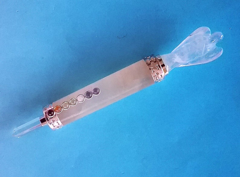 May include: A crystal wand featuring a carved angel at one end and a clear crystal point at the other. The wand has a light green body with seven coloured stones and silver-coloured metal accents. The background is a solid blue.
