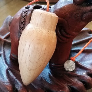 Puede incluir: Un péndulo de madera clara con una cuerda naranja y una pequeña cuenta transparente. El péndulo descansa sobre una base de madera tallada de color marrón oscuro. La base tiene un diseño decorativo.