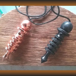 May include: Two pendulums with black cords rest on a wooden surface. One is copper-colored, the other is black. Both have a rounded top and a conical bottom with a series of stacked rings. The copper pendulum has a shiny finish.