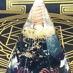May include: A clear pyramid-shaped orgone device with a pointed top. The pyramid contains layers of black and gold materials, with a copper coil at the base. The background is a dark fabric with gold geometric patterns.