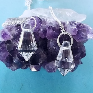 Puede incluir: Dos péndulos de cristal transparente con cadenas plateadas que cuelgan de un grupo de cristales de amatista sobre un fondo azul.