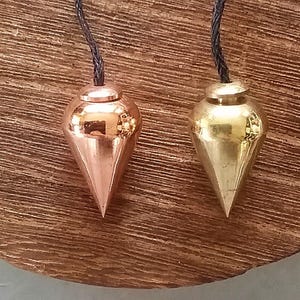 Pode incluir: Dois fios de prumo metálicos, um cor de cobre e outro dourado, pendurados por cordões pretos. Os fios de prumo têm formato de gota com a parte inferior pontiaguda. Estão sobre uma superfície de madeira castanha texturizada.