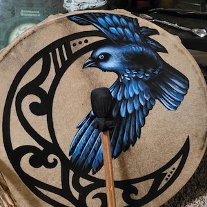 May include: A round tan drum featuring a hand-painted black crescent moon design and a large blue raven in flight. A wooden mallet with a black fabric-covered head rests vertically across the center of the drum face, partially obscuring the bird.