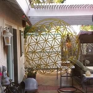 May include: A yellow metal flower of life mandala hanging in a backyard patio. The mandala is a geometric pattern with overlapping circles.