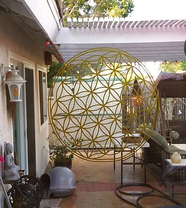 May include: A yellow metal flower of life mandala hanging in a backyard patio. The mandala is a geometric pattern with overlapping circles.