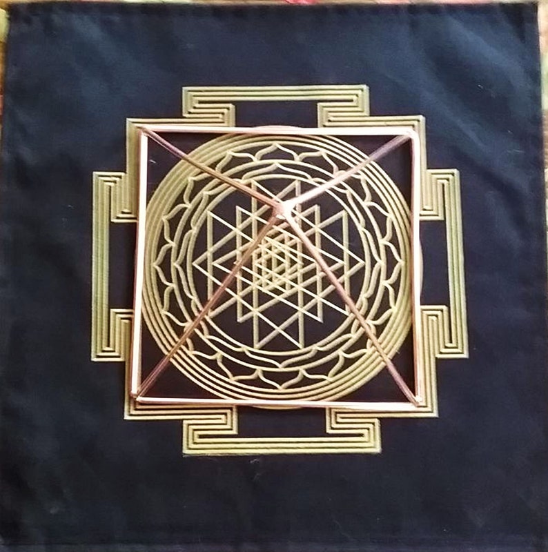 K&ouml;nnte beinhalten: Eine Pyramide aus Kupferdraht liegt auf einem schwarzen Stoffhintergrund mit einem goldenen geometrischen Muster. Die Pyramide ist in einem quadratischen Rahmen zentriert und von einem kreisf&ouml;rmigen Muster umgeben.