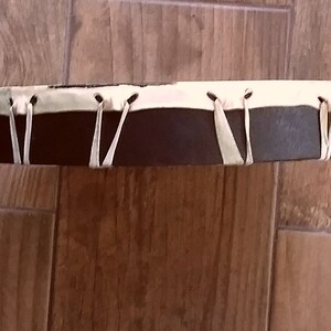 May include: A circular frame drum featuring a dark brown wooden shell and a light cream-colored drumhead. The drumhead is secured to the shell with thin, light-colored leather lacing that wraps around the perimeter in a V-shaped pattern.