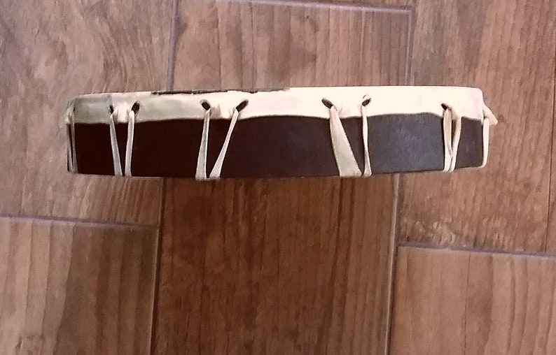 May include: A circular frame drum featuring a dark brown wooden shell and a light cream-colored drumhead. The drumhead is secured to the shell with thin, light-colored leather lacing that wraps around the perimeter in a V-shaped pattern.