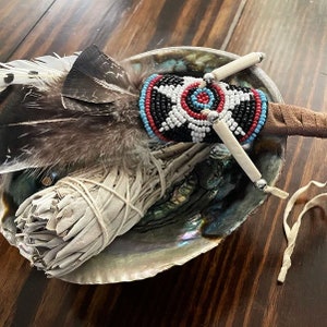 May include: Abalone shell holding a white sage bundle, a beaded wand with feathers, and a decorative wand with bone-like accents. The beaded wand features black, red, white, and blue beads. The feathers are brown and white.