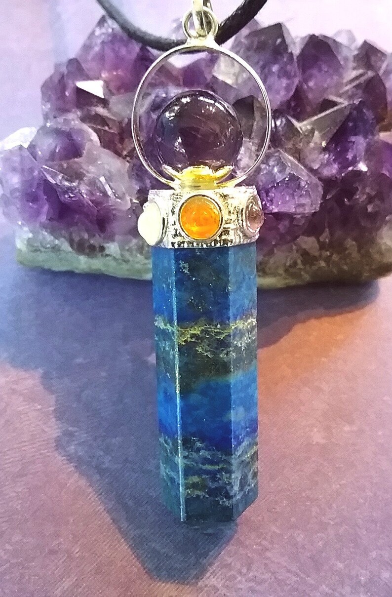 Puede incluir: Un colgante de cristal de lapisl&aacute;zuli azul con un engaste plateado y una esfera de cristal transparente que contiene un cristal de amatista p&uacute;rpura. El colgante est&aacute; rodeado de cristales m&aacute;s peque&ntilde;os de varios colores, incluyendo naranja, rojo y blanco.