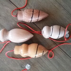 以下が含まれることがあります： 様々な天然木の色合いの木製こまが4つ。それぞれ赤い紐と透明なガラスビーズが付いています。黒い縞模様のあるこまもあります。風化した木製の表面に配置されています。