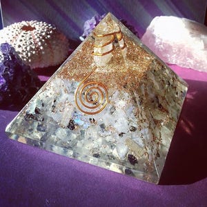 May include: A clear resin pyramid containing embedded white and translucent stone fragments, gold-colored metallic flakes, and a central clear quartz point wrapped in a copper wire coil, featuring a prominent copper spiral on the front face.