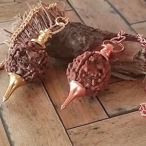 Könnte beinhalten: Zwei braune, runde und strukturierte Pendel mit goldenen und kupfernen Ketten. Die Pendel hängen an einer Holzoberfläche. Das goldene Pendel hat eine goldene Spitze und das kupferne Pendel hat eine kupferne Spitze.