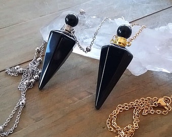 Può includere: Due pendoli di cristallo nero con catene argentate e dorate. Ogni pendolo ha un corpo sfaccettato a forma di cono e una parte superiore sferica. Le catene sono drappeggiate su una superficie di legno, con un cristallo bianco sullo sfondo.