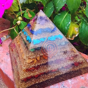 Op de afbeelding: Een zevenlaags kristallen piramide met een koperen draadspiraal in het midden. De piramide is gemaakt van helder hars en bevat verschillende kristallen, waaronder amethist, helder kwarts, citrien, karneol, rode jaspis, turquoise en gouden glitter.