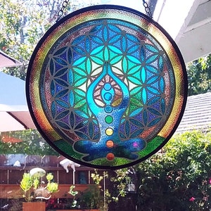 Op de afbeelding: Een rond glas-in-loodraam dat aan een ketting hangt. Het raam heeft een kleurrijk mandala-ontwerp met een mediterend figuur in het midden. De mandala is een geometrisch patroon met een levensbloemontwerp.