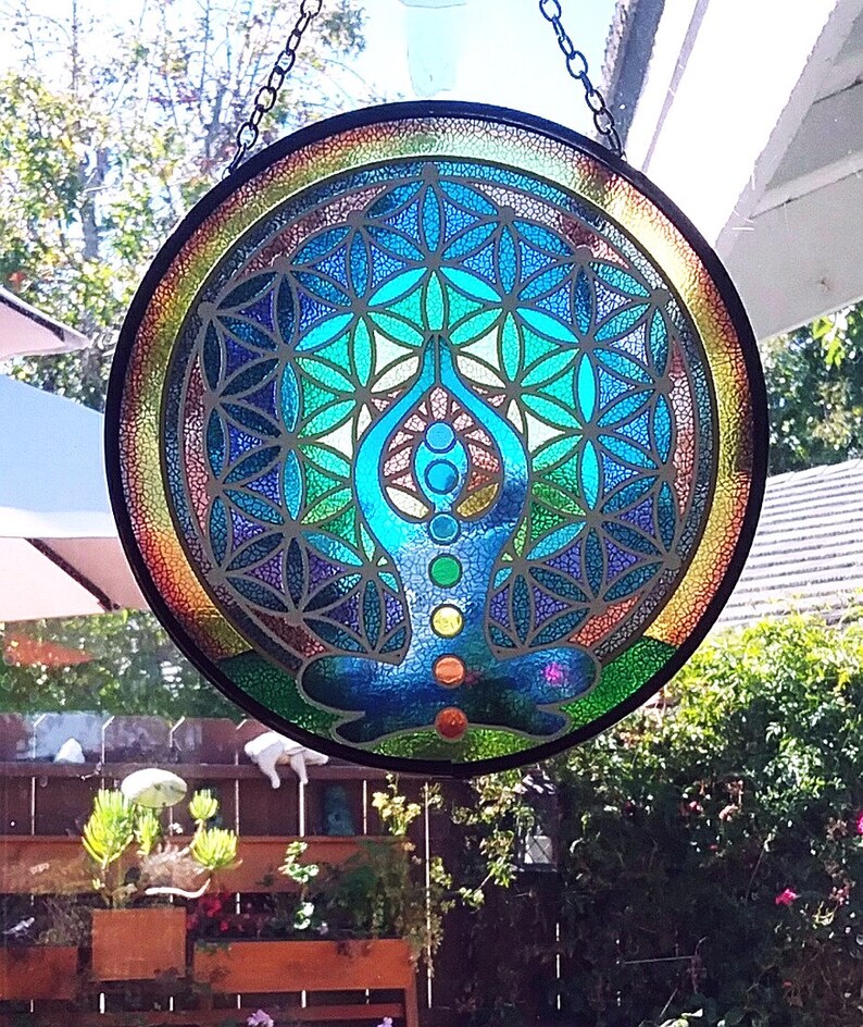 Op de afbeelding: Een rond glas-in-loodraam dat aan een ketting hangt. Het raam heeft een kleurrijk mandala-ontwerp met een mediterend figuur in het midden. De mandala is een geometrisch patroon met een levensbloemontwerp.