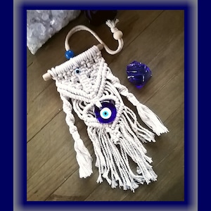 May include: White macrame wall hanging with a blue evil eye charm. The hanging has a wooden dowel at the top and white fringe at the bottom.