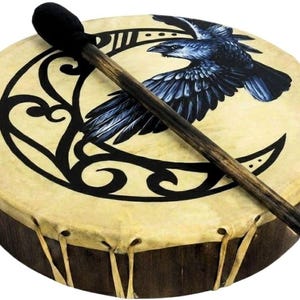 May include: A round frame drum with a light tan drumhead featuring a black graphic of a crescent moon and a flying raven. The drum has a dark wooden frame laced with tan leather strips and includes a wooden mallet with a black felt head.